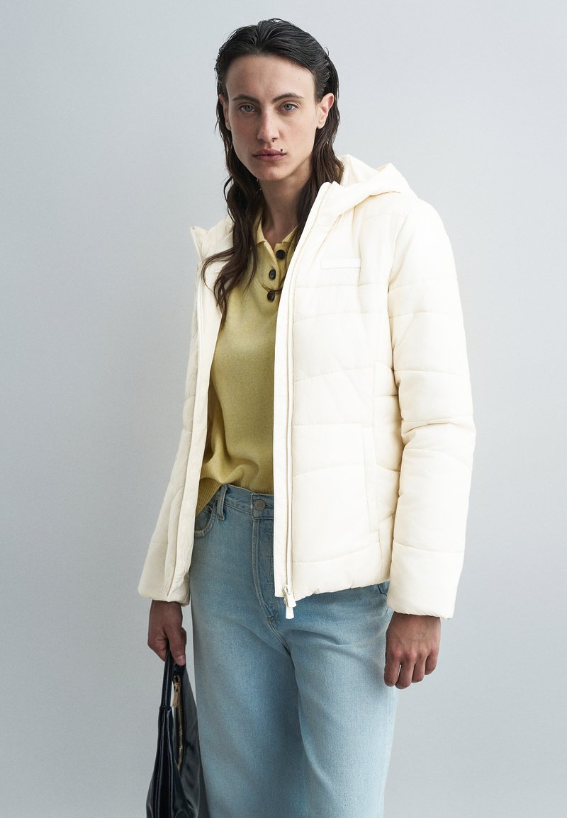 Giacca imbottita di colore crema con cappuccio, dotata di zip, tasche laterali e design trapuntato, indossata sopra un top giallo e jeans blu.