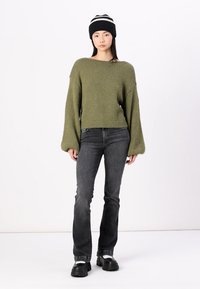 Olivgrüner Strickpullover mit weiten Ärmeln, kombiniert mit schwarzen Jeans und einer schwarzen Strickmütze mit weißen Streifen. Klobige schwarze Stiefel.