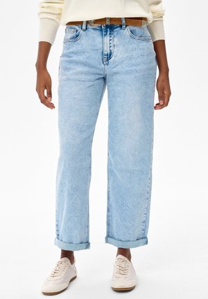 Osoba ubrana w jasnoniebieskie podwinięte dżinsy, beżowy pasek, kremowy sweter i białe trampki stojąca na białym tle.