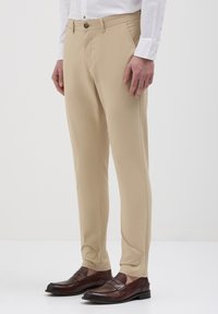 Pantaloni beige su misura con vestibilità dritta, realizzati in tessuto liscio, con chiusura a bottone e tasche laterali, abbinati a mocassini marroni scuri.