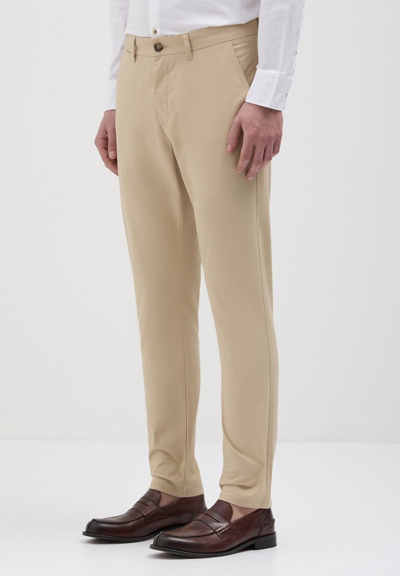 Pantaloni beige su misura con vestibilità dritta, realizzati in tessuto liscio, con chiusura a bottone e tasche laterali, abbinati a mocassini marroni scuri.