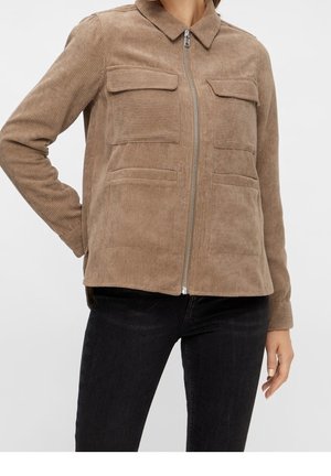 Kvinde iført en beige kordfløjls-zipjakke med bryst- og taljelommer og sorte tætsiddende jeans.
