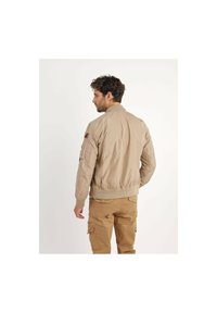 Veste bomber beige avec des poignets et une taille côtelés; dotée d'une fermeture éclair à l'avant, de poches latérales et de patchs sur les manches ; texture de tissu lisse.