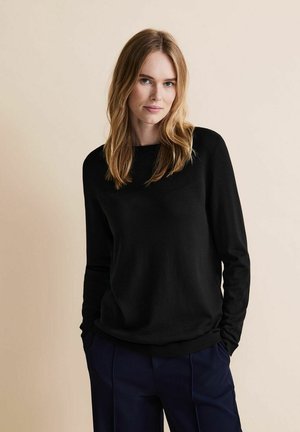 Pull en maille noir avec des manches longues, un col rond et un ourlet côtelé. Tissu texturé, associé à un pantalon bleu ajusté.