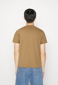 Brązowy bawełniany T-shirt z okrągłym dekoltem i krótkimi rękawami, widoczny z tyłu. Połączony z jasnoniebieskimi dżinsami. Prosty, jednolity design.