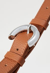 Ceinture en cuir brun avec une surface texturée, une boucle en métal argenté brillant et une bride pour ajuster la taille.