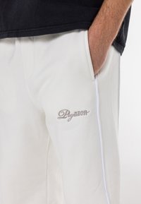 White sweatpants feature a logo embroidered in beige, side stripes in white, and a relaxed fit with pockets. Material appears soft and textured.