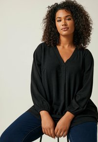 Schwarze, leichte Bluse mit V-Ausschnitt, langen Ärmeln und gerafften Manschetten; kombiniert mit blauer Jeans. Glatte Textur, lockere Passform.