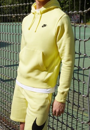 Homme portant un sweat à capuche Nike jaune et un short assorti, debout à côté d'un filet de sport sur un terrain extérieur.