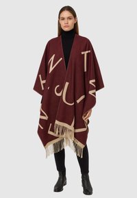 Poncho marrone con grandi lettere crema, orlo a frange e design aperto. Indossato sopra un dolcevita nero, con pantaloni neri aderenti e stivali.