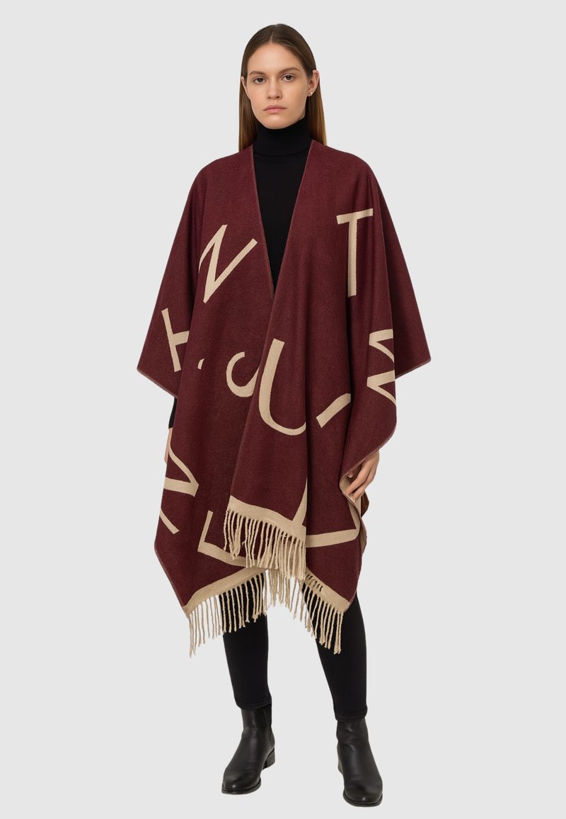 Poncho marrone con grandi lettere crema, orlo a frange e design aperto. Indossato sopra un dolcevita nero, con pantaloni neri aderenti e stivali.