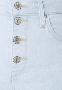 Falda de mezclilla azul claro con cinco botones de metal con grabado de la marca, costuras amarillas en contraste y un primer plano de la textura de la tela.