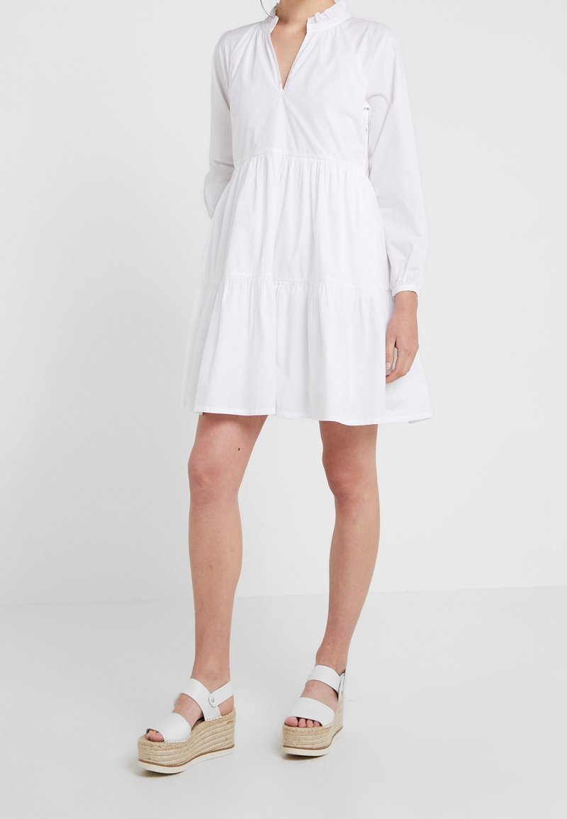 Bílé bavlněné šaty s dlouhými rukávy, véčkovým výstřihem a volánkovým límcem. Mají sukni s volány a volný střih. Doplněné o bílé platformové sandály.