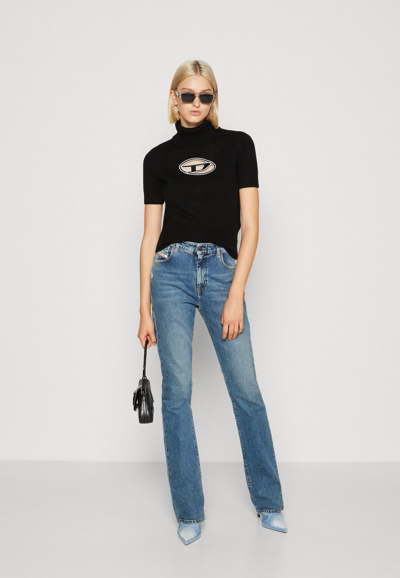 Schwarzes gestreiftes kurzärmliges Rollkragenoberteil mit beigem Logo, kombiniert mit hellblauen Schlagjeans. Das Model hält eine kleine schwarze Handtasche und trägt eine Sonnenbrille.