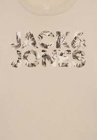 Μπεζ T-shirt με στρογγυλή γραμμή λαιμού που φέρει το "JACK & JONES" με έντονα γράμματα, καλυμμένα με ασπρόμαυρο τροπικό φυλλώδες σχέδιο.