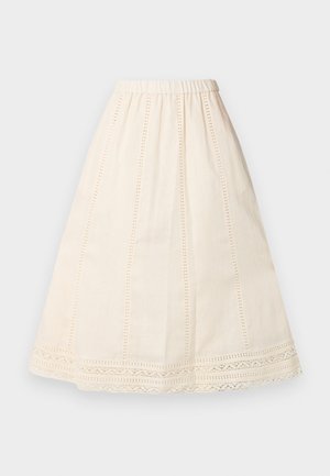 Kobieta stojąca z rękami w kieszeniach, ubrana w biały, drapowany top z krótkim rękawem, kremową spódnicę midi z koronkowymi detalami oraz ciemnobrązowe buty Mary Jane.