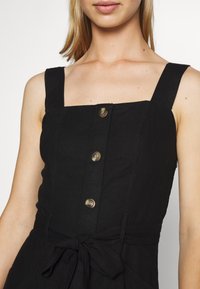 Svart ärmlös klänning med fyrkantig halsringning, tre knappar fram och knytning i midjan. Tyget verkar ha en strukturerad och något texturerad yta.