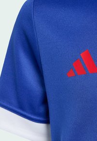 Plava sportska majica s teksturiranim materijalom, bijelim naglascima na rukavima i istaknutim crvenim Adidas logotipom na lijevoj strani.