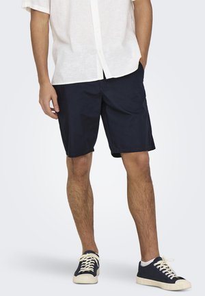 Hombre vestido con camisa blanca de manga corta con botones, pantalones cortos azul marino hasta la rodilla y zapatillas azul marino con cordones y suelas blancas.