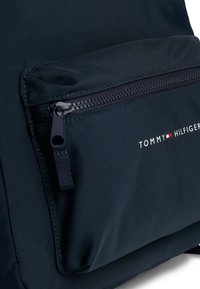 Marinblå ryggsäck i texturerad nylon, med en ficka fram som stängs med dragkedja och en framträdande vit logotyp på sidan.