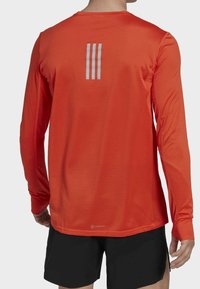 T-shirt de sport à manches longues de couleur orange vif, avec un tissu texturé et trois bandes réfléchissantes au dos. Shorts noirs présentés en dessous.