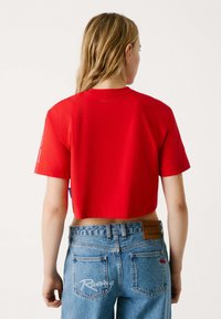 T-shirt croppato rosso in un tessuto rosso brillante. Maniche corte, scollatura rotonda e un sottile dettaglio del logo sul retro. Indossato con jeansazzurri chiari.