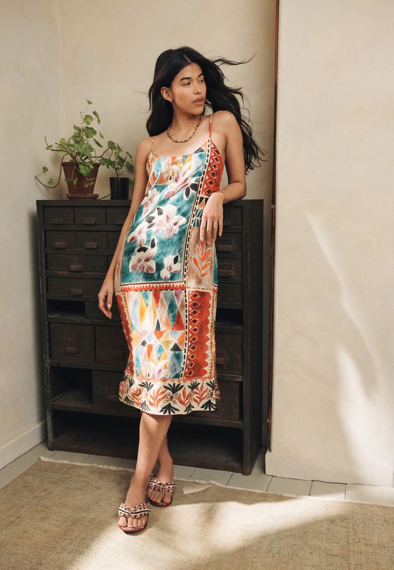 Femme aux longs cheveux foncés portant une robe colorée à motifs floraux et géométriques et des sandales, debout et appuyée contre une armoire en bois foncé.
