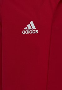 Veste de sport rouge en tissu lisse, avec un logo Adidas blanc sur la poitrine. Design simple avec des détails de couture minimalistes.