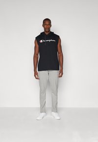 Schwarze ärmellose Kapuzenjacke mit weißem "Champion"-Logo, kombiniert mit hellgrauen Sweatpants und weißen Sneakers. Schlichte, legere Sportbekleidung.