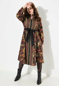 Abito a maniche lunghe con motivo paisley in colori ricchi, cintura nera e scollo con bottoni. Abbinato a stivaletti neri alla caviglia.