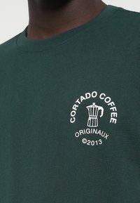 Tume roheline puuvillane T-särk, millel on valge graafika kohvikeetjast koos tekstiga "CORTADO COFFEE ORIGINALAUX ©2013" ümber.