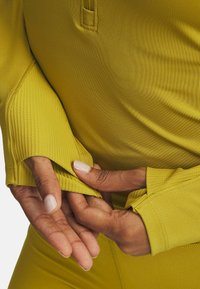 Close-up van handen die de duimopening manchet van een langärmelige, mosterdgele sporttop met een aansluitende pasvorm en bijpassende leggings aanpassen.