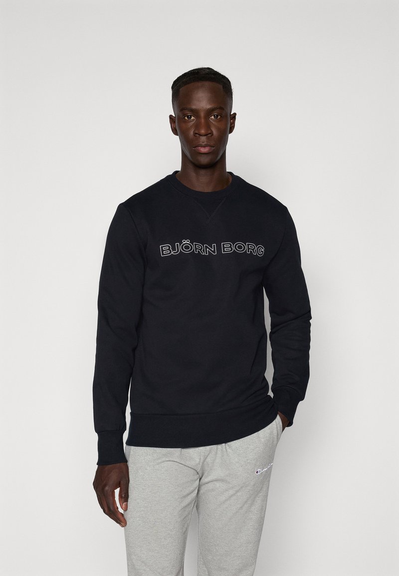Μαύρο φούτερ με το λογότυπο "BJÖRN BORG" σε λευκό, στρογγυλή λαιμόκοψη, μακριά μανίκια, κατασκευασμένο από μαλακό ύφασμα. Συνδυασμένο με γκρι φόρμες.