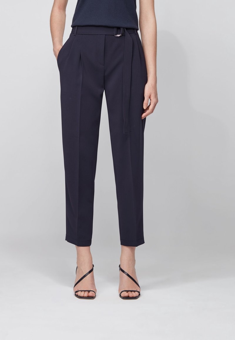Femme portant un pantalon taille haute bleu marine tailleur avec ceinture, des sandales noires à talons hauts à brides, et un haut bleu marine, debout devant un fond uni.
