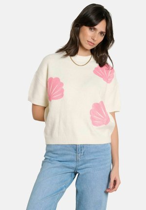 Frau trägt einen weißen Pullover mit kurzen Ärmeln und rosa Muschelmuster, blaue Jeans und eine goldene Halskette vor einem einfarbigen Hintergrund.