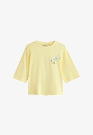 Světle žluté krátké tričko s kreslenými postavičkami Snoopyho a Woodstocka u potisku pampelišky na levé straně hrudi.