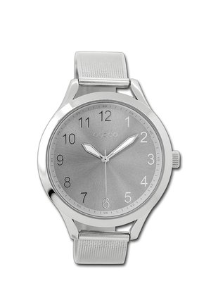 Montre argentée avec un bracelet métallique, cadran rond, fond gris, chiffres arabes et deux aiguilles argentées. Logo de la marque visible en haut.