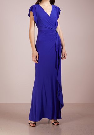 Woman wearing a floor-length royal blue dress with flutter sleeves, V-neck, side drape, and jeweled waist detail, paired with black heels.