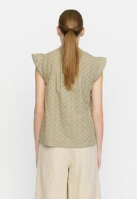 Kaki ärmlös topp med fladdrande ärmar, prydd med broderade ögondetaljer och rund halsringning, matchad med ljusbeiga byxor.