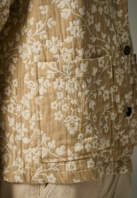 Jachetă bej matlasată cu broderie florală albă, un buzunar pe piept și nasturi negri. Textura este moale, iar materialul pare ușor.