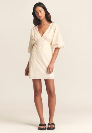 Korte mouw, crèmekleurige jurk met een diepe V-hals, geraffineerde taille en een oogpatroon. De stof is lichtgewicht en lichtjes gestructureerd.