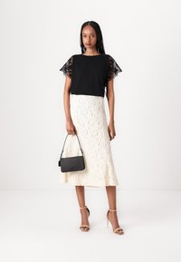 Top negro con mangas de encaje, falda blanca texturizada y bolso negro. Unas sandalias de tiras en colores neutros completan el conjunto.