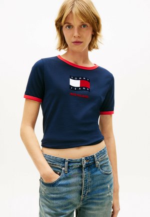 Junge Person mit kurzem blondem Haar, die ein marineblaues kurz geschnittenes Oberteil mit rotem Rand und einem Tommy Jeans-Logo trägt, kombiniert mit blauen Jeans.