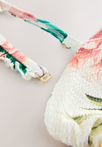 Pisano cvetlični top iz teksturirane belega tkanine, z nagubanim trakovi v roza in zeleni barvi ter z zlatim okovjem za pritrditev trakov.