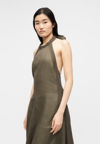 Γυναίκα με αμάνικο ελαιί πράσινο φόρεμα με πλεκτές λεπτομέρειες, στέκεται μπροστά σε απλό ανοιχτό γκρι φόντο, κοιτάζοντας εμπρός με ουδέτερη έκφραση.