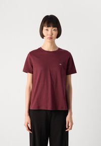 Young woman with short black hair wearing a burgundy short-sleeve t-shirt and black pants, standing against a plain light background.