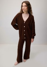 Brown cable-knit cardigan with a v-neck and large buttons, paired with matching flared pants. Soft texture, worn with light pink shoes.