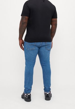 Homme avec des tatouages portant un t-shirt noir, un jean bleu et des baskets noires, debout dos à un fond blanc uni.
