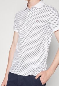 Vit polo-t-shirt med ett mönster av små svarta ovaler, korta ärmar och en klassisk krage. Diskret logotyp på bröstet.