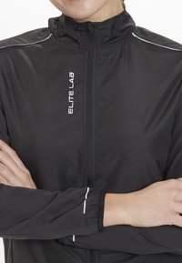 Sort letvægtsjakke med højt krave, fuld lynlås og reflekterende detaljer. Har elastiske manchetter og "ELITE LAB" branding på fronten.